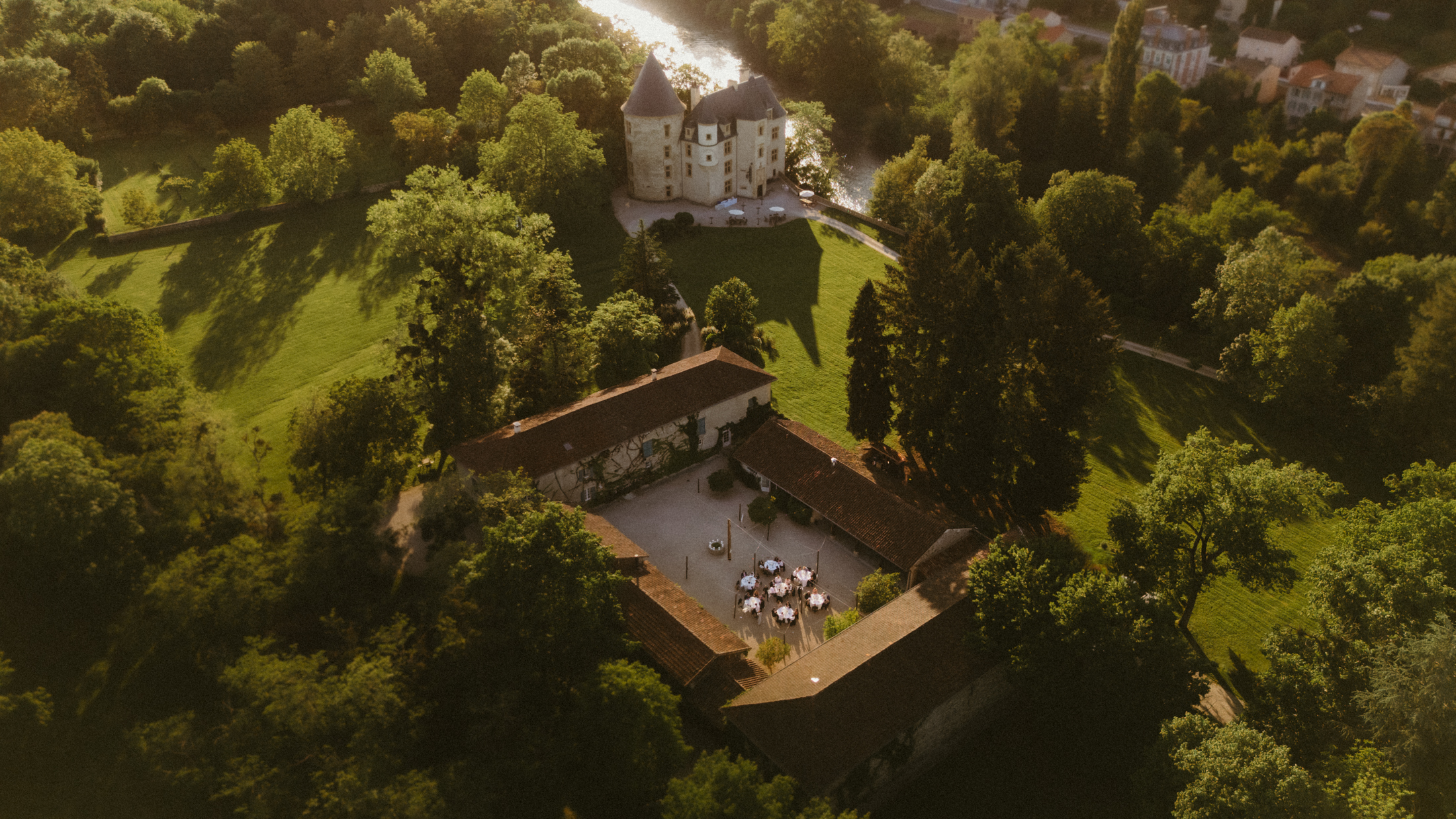 Aerial photo of a French château at sunset, surrounded by green forest, with wedding dinner tables arranged in the courtyard.